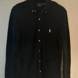 Svart meshskjorta från Polo Ralph Lauren - Svart långärmad skjorta från Polo Ralph Lauren i featherweight mesh. Klassisk button-down krage, vita knappar och ikonisk broderad logga på bröstet. Skjortan har en normal passform och är tillverkad i lätt bomullsmaterial som andas.