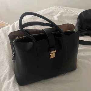 Svart handväska med gulddetaljer - Stilren svart handväska med två rejäla handtag och snygga gulddetaljer på låset. Väskan har en praktisk dragkedja upptill och är tillverkad i skinnimitation. Insidan är rymlig och fodrad i beige ton, perfekt för att få plats med allt du behöver.