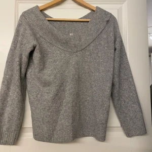 Grå stickad tröja med v-ringning - Mysig grå stickad tröja med bred v-ringning och långa ärmar. Tröjan har ribbade muddar vid ärmslut och nederkant, perfekt för kyliga dagar. Enkel och stilren design som passar till det mesta.