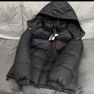 Svart pufferjacka från Polo Ralph Lauren - Svart pufferjacka från Polo Ralph Lauren med huva och dragkedja framtill. Jackan har en röd broderad logga på bröstet och elastiska muddar vid ärmsluten. Perfekt för kalla dagar och riktigt snygg streetstil.