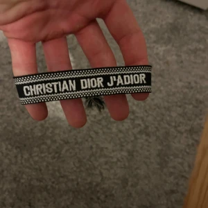 Christian Dior J'Adior armband - Snyggt armband från Christian Dior med texten 'J'Adior' broderad i vitt på svart bakgrund. Armbandet har ett randigt mönster och knytning med tofsar i ändarna. Perfekt accessoar för att lyfta din outfit med en lyxig touch.