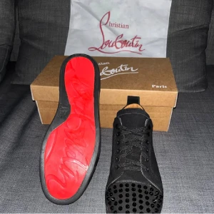 Christian Louboutin svarta sneakers med nitar - Svarta sneakers från Christian Louboutin med klassisk röd sula och svarta nitar på tån. Skorna har snörning och är tillverkade i mocka med detaljer i skinn. Snygg och edgy design som sticker ut, perfekt för dig som gillar statement pieces.