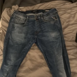 Replay Anbass blå jeans, storlek 34 - Snygga Replay Anbass jeans i blå tvätt med slitningar och klassiska detaljer. Modellen har smal passform, fem fickor och dragkedja. Tuffa kontrastsömmar och Replay-logga på fickan ger extra stilpoäng. Perfekt för dig som gillar en modern look.