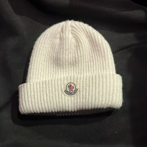 Moncler mössa - Snygg vit ribbstickad mössa från Moncler med klassisk uppvikt kant och broderad logotyp framtill. Tillverkad i mjukt material som håller dig varm. Perfekt mössa till vintern.