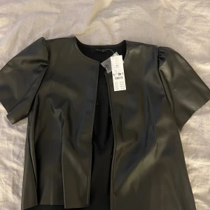 Svart blus från Gina Tricot XS - Säljer en svart blud från Gina Tricot i storlek XS. Blusen har korta ärmar med lätt puff och rund halsringning. Den är tillverkad i syntetmaterial med en slät, glansig finish och har en rak, något boxig passform. Perfekt för dig som gillar trendiga statement-plagg. Helt ny, aldrig använd