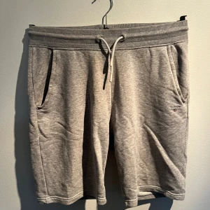 Grå mjukisshorts från Gant - Säljer ett par grå mjukisshorts från Gant med ribbad midja och snörning framtill. Shortsen har två sidofickor och en liten broderad logga på vänster ben. Perfekta för chill eller träning, tillverkade i mjuk bomullsmix.