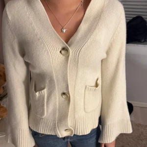 Krämvit kofta från Stylein XS - Superfin krämvit kofta från Stylein i storlek XS. Koftan har v-ringning, stora knappar framtill och två små fickor. Den är ribbstickad vid ärmslut och nederkant, vilket ger en snygg och stilren look. Perfekt att slänga över en t-shirt eller linne.