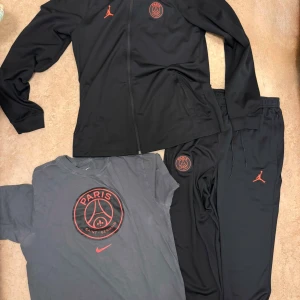 Svart PSG x Jordan vindjacka med huva - Svart vindjacka från Jordan x PSG med huva, hel dragkedja och röda loggor på bröstet. Jackan har två fickor med dragkedja och är tillverkad i lätt polyester. Perfekt för dig som gillar sportig och clean streetstyle.