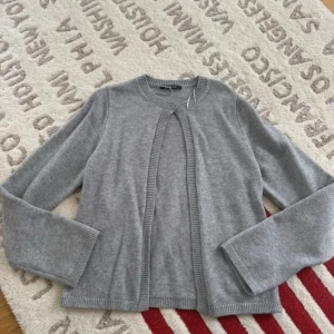 Grå kofta från Gina Tricot - Mysig grå kofta från Gina Tricot med rund halsringning och en knapp framtill. Koftan är stickad i mjukt material och har långa ärmar samt ribbade kanter vid hals, ärmslut och nederkant. Perfekt att slänga över en t-shirt eller linne.