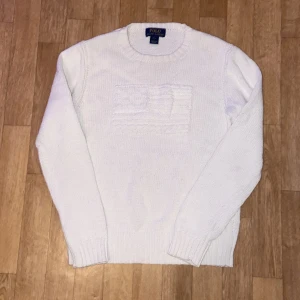 Vit stickad tröja från Polo Ralph Lauren - Vit stickad tröja från Polo Ralph Lauren med rund halsringning och ett diskret stickat flaggmönster. Tröjan har ribbade muddar vid ärmslut och nederkant, perfekt för lager-på-lager under kyliga dagar.