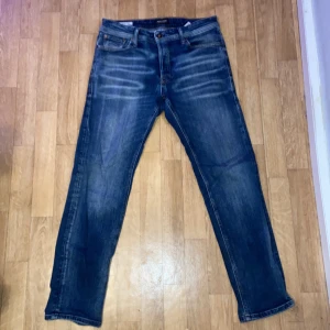 Blå jeans från Jack & Jones, storlek 31/30 - Snygga blå jeans från Jack & Jones med klassisk femficksdesign med en fin wash framtill. Modellen har normal passform och raka ben, tillverkade i jeansmaterial med bekväm midja. Perfekt för dig som gillar stilrena och tidlösa jeans. Dom har en liten slitning där bak pågrund av att lappen har gått av där bak men annars inga slitningar.