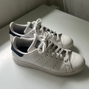 Adidas Stan Smith vita sneakers - (SKICKAS NYTVÄTTADE) Klassiska Adidas Stan Smith sneakers i vitt skinn med mörkblå detaljer på hälen och tungan. Skorna har rund tå, snörning och platt sula med ikonisk Adidas-logga. Perfekta för en clean och tidlös stil.
