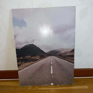 Väggtavla med landskapsfoto av väg - Stilren väggtavla med motiv av en landsväg mot berg i dimma. Tavlan har synligt slitage och skador på nederkanten, men motivet är intakt. Perfekt för att skapa en känsla av frihet i hemmet. Utrustad med upphängningsbeslag på baksidan.
