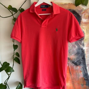 Röd pikétröja Polo Ralph Lauren - Snygg röd pikétröja från Polo Ralph Lauren, modell The Earth Polo Custom Slim Fit. Klassisk krage, kort ärm och grönt broderat logomärke på bröstet. Perfekt för en clean och sportig look.