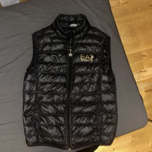 Svart dunväst från EA7 Emporio Armani - Svart dunväst från EA7 Emporio Armani med guldfärgad logga på bröstet. Västen har quiltad design, hög krage och dragkedja framtill. Perfekt för lager-på-lager och har en snygg, sportig look som funkar till många stilar.