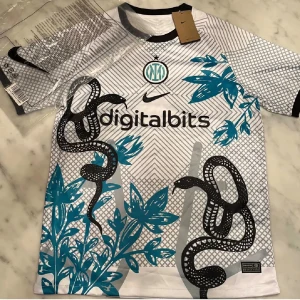 Inter Milan x Nike vit fotbollströja - Säljer en Inter Milan fotbollströja från Nike i vit med svart och turkos detaljer. Tröjan har korta ärmar, coolt grafiskt mönster med ormar och blommor, samt klubbmärke och sponsortryck på bröstet. Perfekt för dig som vill sticka ut på planen.