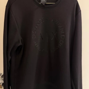 Svart sweatshirt från Armani Exchange - Svart sweatshirt från Armani Exchange i storlek L. Tröjan har ett stilrent, ton-i-ton tryck med A|X-logga och text på bröstet. Rund halsringning och långa ärmar. Perfekt för dig som gillar minimalistisk design och vill ha en snygg, enkel look.