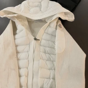Vit dunjacka från Moncler med huva - Säljer en snygg vit dunjacka från Moncler med ribbade ärmar och quiltad front. Jackan har en stor huva, dragkedja och coolt Moncler-märke på ärmen. Insidan har ett färgglatt serietryck. Perfekt för dig som vill ha en clean och exklusiv look.oanvänd finns alla storlekar 