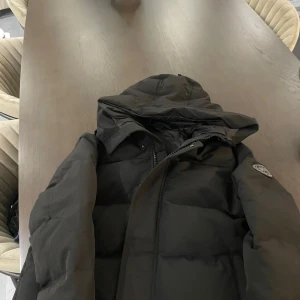 Svart pufferjacka från Canada Goose - Svart pufferjacka från Canada Goose med huva och snyggt märke på ärmen. Jackan har dragkedja och tryckknappar framtill, samt två praktiska fickor. Perfekt för kalla dagar och har en clean, modern look som funkar till allt. Den är oanvänd fick den i present men den satt inte bra har även en likadan i storlek large
