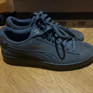 Mörkgråa Puma sneakers i mocka - Säljer ett par sparsamt använda mörkgråa Puma sneakers i mocka med klassisk siluett och guldfärgad Puma-logga på sidan. Skorna har snörning och platt sula, samt diskreta detaljer som ger en clean look. Perfekta för dig som gillar stilrena och bekväma sneakers. Har en missfärgning på skorsnöret, har inte testat att tvätta bort de.