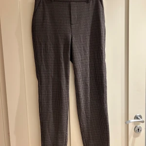 Rutig kostymbyxa från Vero Moda - Snygga kostymbyxor från Vero Moda i storlek S/32 med ett diskret rutigt mönster i mörkgrå och beige toner. Byxorna har elastisk midja bak, bälteshällor och raka ben. Perfekt för dig som gillar klassisk stil med en modern twist.