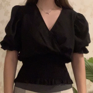 Svart omlottblus med puffärm - Säljer en svart blus med omlott-design och v-ringning. Toppen har korta puffärmar och en smockad midja. 