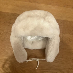 Vit pälsmössa från H&M M/L - Säljer en fluffig vit pälsmössa från H&M i storlek M/L. Mössan har öronlappar och snörning under hakan för extra värme och stil. Insidan är fodrad med glansigt tyg som känns mjukt mot huden. Perfekt för kalla vinterdagar och ger en cool vibe.