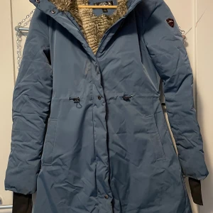Blå parkajacka från Platinum XS - Säljer en blå parkajacka från Platinum i storlek XS. Jackan har en mysig fuskpäls runt kragen, långa ärmar och praktiska fickor. Den stängs med dragkedja och knappar, och har snörning i midjan för snygg passform. Perfekt för kalla dagar! NYPRIS 2000