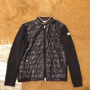 Svart quiltad cardigan jacka Moncler - Svart cardigan jacka från Moncler med quiltad front och släta ärmar. Jackan har ribbad krage med röd och vit rand, dragkedja framtill och Moncler-logga på ärmen. Materialet är en mix av polyester och bomull, perfekt för dig som gillar stilren design.