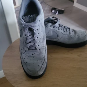 Grå Nike Air Force 1 sneakers mocka - Säljer ett par grå Nike Air Force 1 sneakers i mocka med svart platt sula. Klassisk rund tå och snörning framtill. Snyggt ton-i-ton utseende med diskret Nike-logga på sidan och tungan. Perfekt för dig som gillar stilrena och tidlösa sneakers.