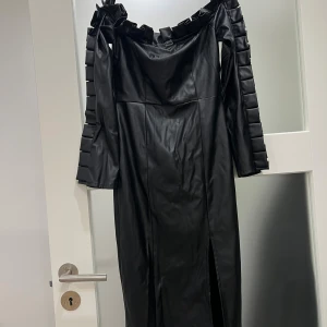 Svart lång klänning med volang - Säljer en svart lång klänning i skinnimitation med offshoulder och volangdetaljer längs halsringningen och ärmarna. Klänningen har långa ärmar och en snygg slits framtill som ger extra edge. Perfekt för dig som vill sticka ut med en cool och unik look.