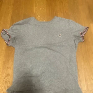 Grå Moncler t-shirt med logga - Snygg grå t-shirt från Moncler med klassisk logga på bröstfickan. T-shirten har korta ärmar med röd, vit och marinblå randig kant. Tillverkad i mjuk bomull och har en normal passform. Perfekt för en clean och stilren look.