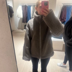 Brun teddyjacka med dragkedja - Säljer en brun teddyjacka med hög krage och breda ärmar. Jackan har en grov dragkedja framtill och två stora fickor. Materialet är mjukt och fluffigt, perfekt för kyliga dagar. Passformen är loose och jackan har en avslappnad vibe.