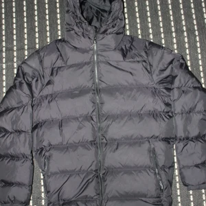 Svart pufferjacka från Neomondo XL - Svart pufferjacka från Neomondo i storlek XL. Jackan har huva med snörning, två sidofickor med dragkedja och quiltad design. Perfekt för kalla dagar med sin fluffiga och isolerande känsla. Stängs med dragkedja framtill och har långärmade ärmar.