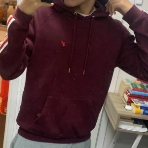 Vinröd hoodie med ränder på ärmarna - Säljer en vinröd hoodie från Lyle & Scott med klassisk huva och snörning. Tröjan har vita, orange och röda ränder längs ärmarna samt en liten röd logga på bröstet. Stor ficka framtill och ribbade muddar. Perfekt för chill dagar.