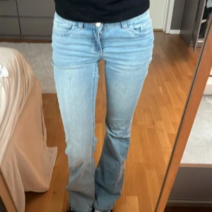 Nelly ljusblå bootcut jeans - Säljer ett par ljusblå jeans från Nelly med bootcut passform och låg midja. Jeansen har klassiska fem fickor, snygga röda sömmar på bakfickorna och dubbla knappar fram. Perfekt för dig som gillar en avslappnad och trendig stil.