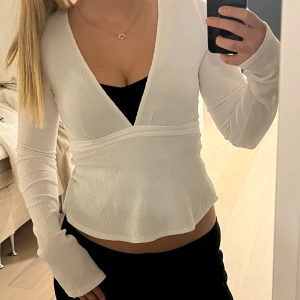Vit topp med knyt och v-ringning - Säljer en vit topp med lång ärm och djup v-ringning. Toppen har ett snyggt knytband i midjan som kan stylas på olika sätt. Materialet är lätt och har en diskret struktur som ger en cool vibe. Perfekt att matcha med jeans eller kjol för en fräsch look.