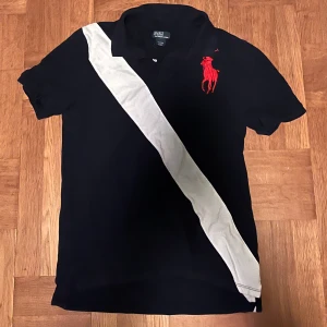 Mörkblå Polo Ralph lauren cheif keef shirt - Snygg mörkblå t-shirt från Polo Ralph Lauren med klassisk krage, korta ärmar och en vit diagonal rand fram och bak. Stor röd broderad logga på bröstet. Tillverkad i mjuk bomull, perfekt för en 2010 cheif keef look 
