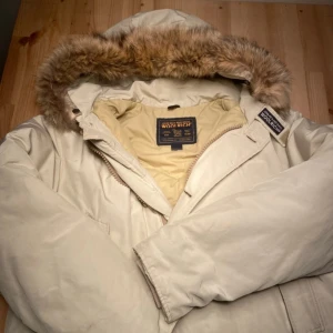 Beige dunjacka med päls från Woolrich strl M - Säljer en beige dunjacka från Woolrich med fluffig päls på huvan och snygga detaljer. Jackan har dragkedja och långa ärmar, perfekt för kalla vinterdagar. Insidan är ljusgul och jackan har en klassisk, rymlig passform. Märket syns tydligt på både insidan och utsidan. Storlek M.