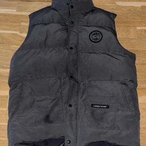 Svart dunväst från Canada Goose - Snygg svart dunväst från Canada Goose med hög krage och två stora fickor framtill. Västen har ett broderat logomärke på bröstet och knäppning med tryckknappar. Perfekt för kalla dagar och har en klassisk, stilren look. Står storlek xxl men den passar L/M då jag själv använt den och är storlek M/L