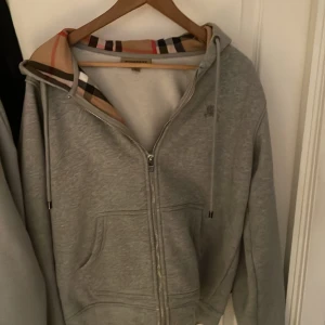 Grå Burberry hoodie med dragkedja - Grå hoodie från Burberry i storlek M med klassiskt rutigt foder i huvan. Tröjan har dragkedja framtill, snörning vid huvan och två fickor. Materialet är mjukt och känns lyxigt, perfekt för dig som gillar designerplagg med ikoniska detaljer.