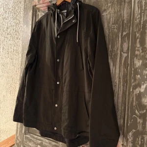 Svart vindjacka Race Marine XL - Svart vindjacka från Race Marine i storlek XL. Jackan har huva med snörning, tryckknappar framtill och två sidofickor. Materialet är lätt och känns sportigt, perfekt för dig som gillar enkel och clean stil. Skandinavisk design med diskreta detaljer.