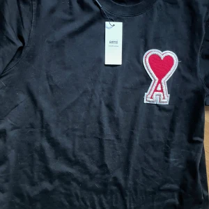 Svart t-shirt från Ami med hjärtdetalj - Svart t-shirt från Ami Alexandre Mattiussi med klassisk passform och rund hals. På bröstet sitter en stor röd hjärtpatch med bokstaven A i vitt. T-shirten är i mjuk bomull och har kortar armar.