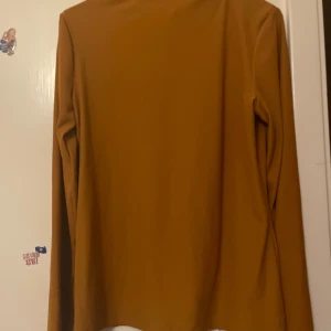 Senapsgul långärmad polotröja - En stilren senapsgul polotröja med lång ärm och lätt ribbad struktur. Tröjan har en enkel, rak passform och en liten ståkrage som ger en clean look. Den är skönt när du har på och den är ny jag har köpt en månad sen