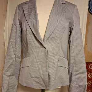 Ljusgrå randig kavaj från H&M - Snygg ljusgrå damkavaj från H&M i storlek 38 med tunna vita ränder. Klassisk passform med slag, två fickor med lock och knäppning framtill. Perfekt för en stilren look. Materialet är bomull, nylon, spandex och polyester. Medföljer 2 extraknappar