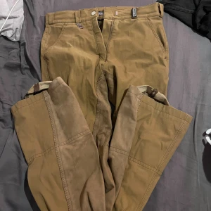 Bruna ridbyxor täckbyxor från Pikcur  - Säljer ett par bruna ridbyxor täckbyxor från Pikcur med förstärkta partier på insidan av benen och praktiska fickor. Byxorna har klassisk passform och är perfekta för utomhusaktiviteter under kyligare dagar.