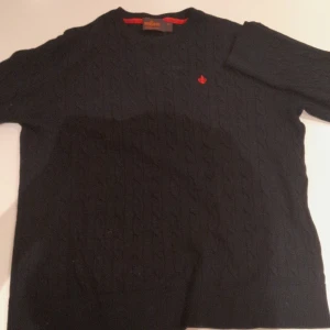 Svart kabelstickad tröja Morris XL - Svart kabelstickad tröja från Morris i 100% merinoull. Klassisk modell med rund halsringning och diskret röd logga broderad på bröstet. Perfekt för dig som gillar stilrena och tidlösa plagg med en snygg twist.