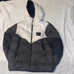 Nike Air Max pufferjacka Storm-FIT M - Nike Air Max pufferjacka i svart och vitt med huva och Storm-FIT-teknologi. Jackan har dragkedja framtill, två dragkedjeförsedda fickor och en snygg Air Max-logga på bröstet. Tillverkad i 100% polyester för att stå emot väder och vind.