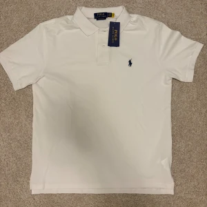 Vit piké från Polo Ralph Lauren - Vit pikétröja från Polo Ralph Lauren i modellen Custom Slim Fit. Klassisk krage, korta ärmar och två knappar framtill. Ikonisk blå broderad logga på bröstet. Tillverkad i mjuk bomull som andas och känns skön mot huden.
