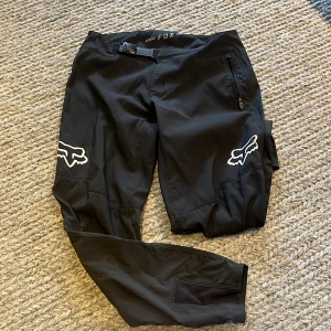 Svarta täckbyxor från Fox Defend - En Fox-storlek Y28 motsvarar ungefär en midja på 71 cm (cirka 28 tum).   Svarta täckbyxor från Fox Defend med vit logga på båda benen. Byxorna har justerbart bälte i midjan, dragkedjeförsedda fickor och är tillverkade i slitstarkt syntetmaterial. Perfekta för dig som gillar att vara aktiv utomhus och vill ha grym stil.